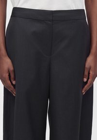 Gros plan d’une personne portant un pantalon noir taille haute à jambes larges, avec une ceinture plate sans attaches visibles.