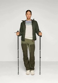 Ženska v zeleni zunanji jakni in hlačah, ki drži črne pohodniške palice, stoji na enobarvni svetli podlagi.