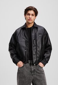 Svart faux skinnjakke med trykknappfront, elastiske mansjetter og en avslappet passform. Kombinert med grå, lett slitte jeans.
