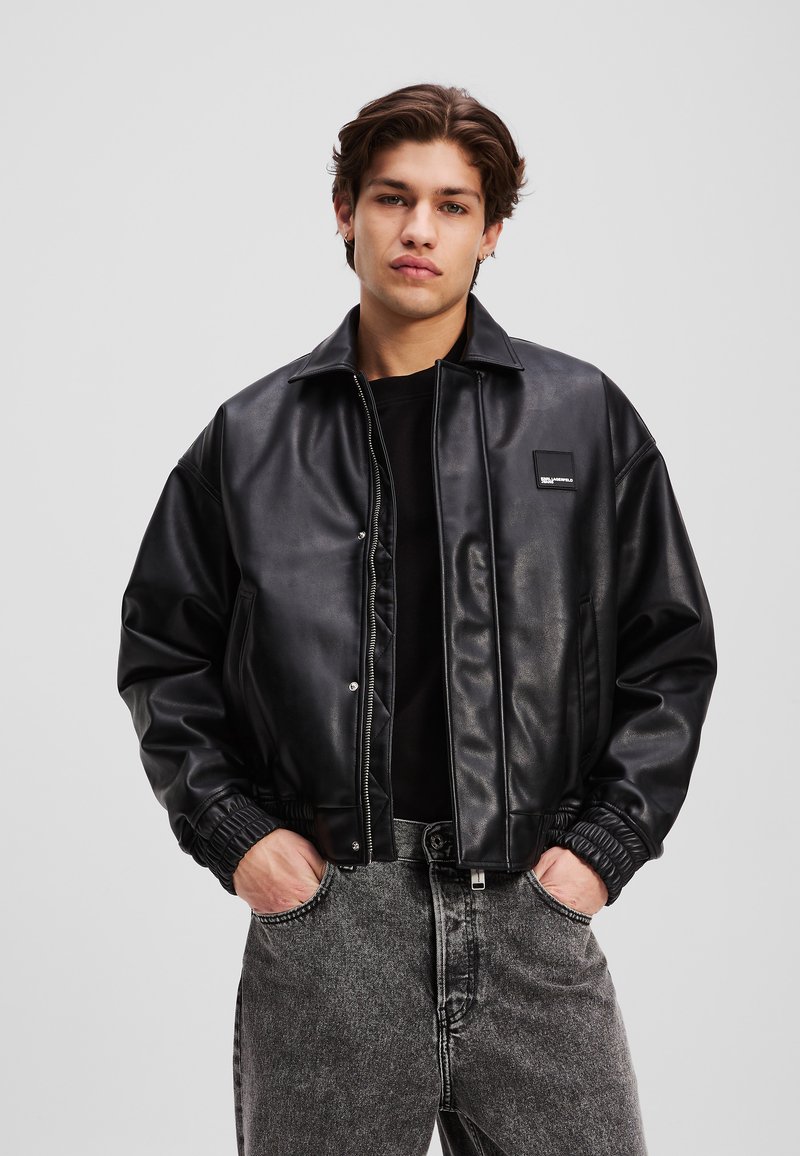 Svart faux skinnjakke med trykknappfront, elastiske mansjetter og en avslappet passform. Kombinert med grå, lett slitte jeans.