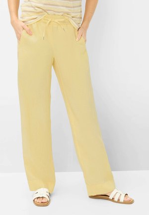 Person trägt lockere gelbe Kordelzughosen, gestreiftes Oberteil und weiße Sandalen, steht mit den Händen in den Taschen vor einfarbigem Hintergrund.