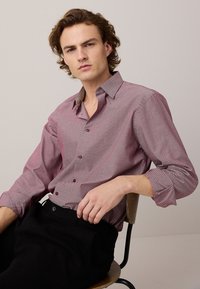 Giovane uomo con capelli mossi seduto, indossa una camicia bordeaux a maniche lunghe con motivo e pantaloni neri, su uno sfondo semplice grigio chiaro.