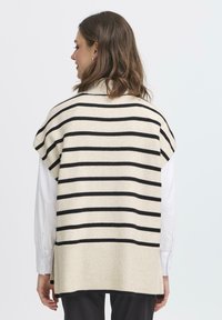 Frau mit schulterlangem braunen Haar, die einen cremefarbigen und schwarzen horizontal gestreiften ärmellosen Strickjäckchen über einem weißen Langarmshirt trägt.