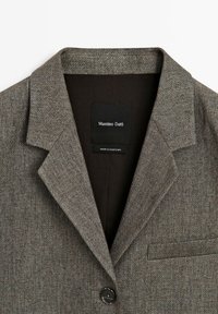 Ruskea teksturoidulla bleiserillä, jossa on viiltoaukolla varustettu kaulus, yksittäinen nappikiinnitys ja rintatasku. Sisämerkissä lukee "Massimo Dutti, Valmistettu Portugalissa."