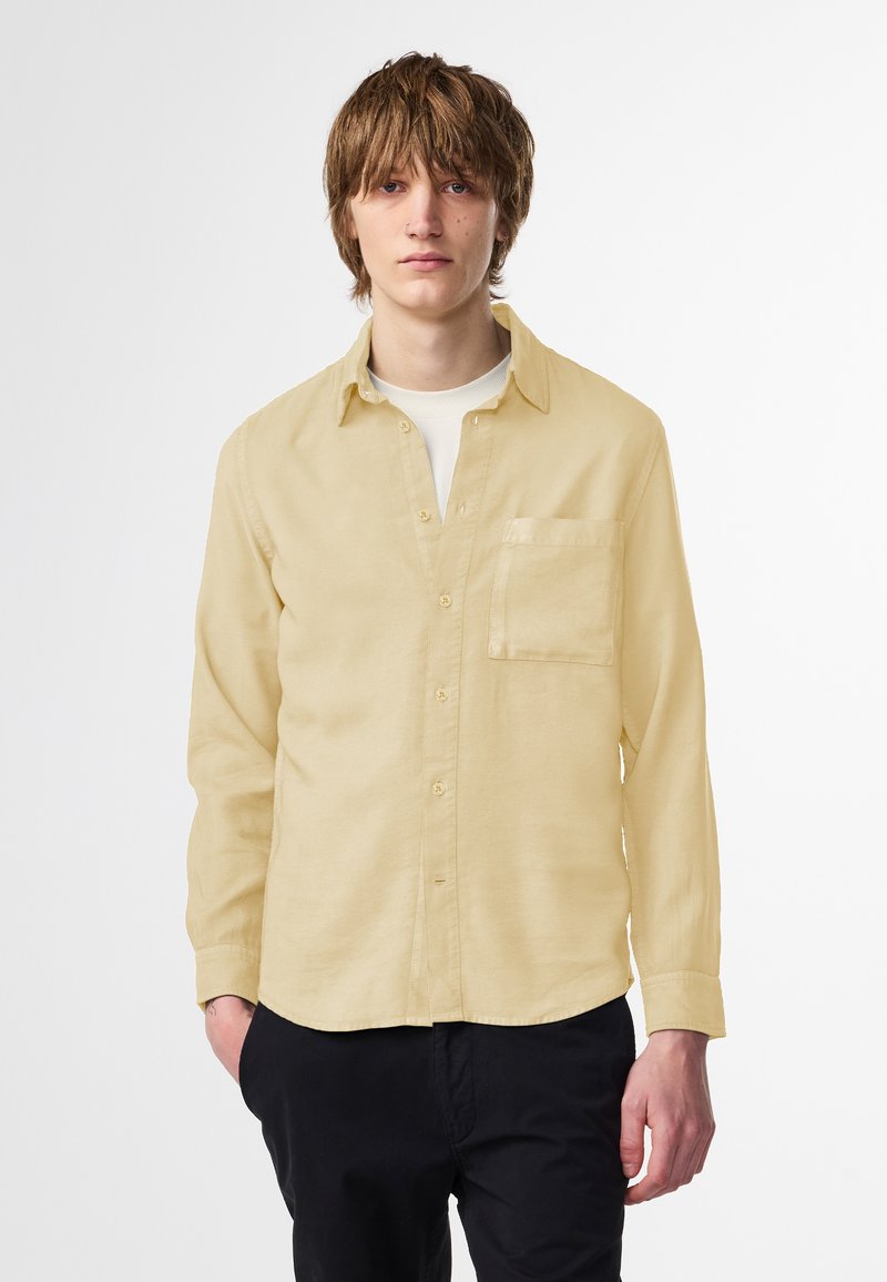 Helles gelbes Hemd mit langen Ärmeln, Brusttasche und lockerer Passform. Aus einem weichen, strukturierten Gewebe gefertigt. Kombiniert mit dunklen Hosen.