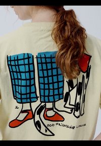 Dos d'une chemise crème avec une illustration de jambes en pantalon à carreaux bleus et chaussures rouges, debout à côté d'un chien noir et blanc. Le texte indique "Amitié canine."