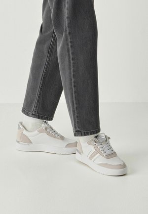 Baskets blanches avec des accents beige clair. Comprennent une tige texturée, un système de laçage et un détail métallique au talon. Portées avec un jean foncé.