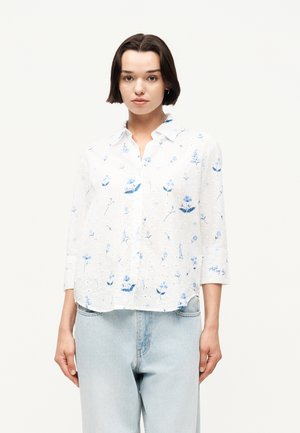 Frau mit kurzen dunklen Haaren, die ein weißes Hemd mit blauen Blumenmustern und hellblaue Jeans trägt, vor einem einfachen Hintergrund stehend.