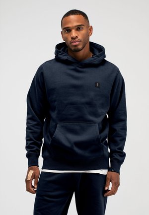 Man draagt een marineblauwe hoodie en bijpassende broek, staat met ontspannen handen, licht verband om linkerhand, neutrale grijze achtergrond.