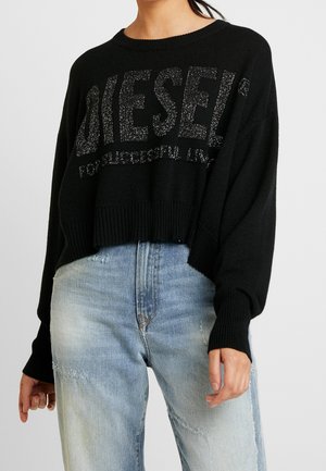 Sort cropped sweater med glitrende "DIESEL" tekst båret med lyseblå højttaljede slidte jeans på en person mod en ensfarvet baggrund.