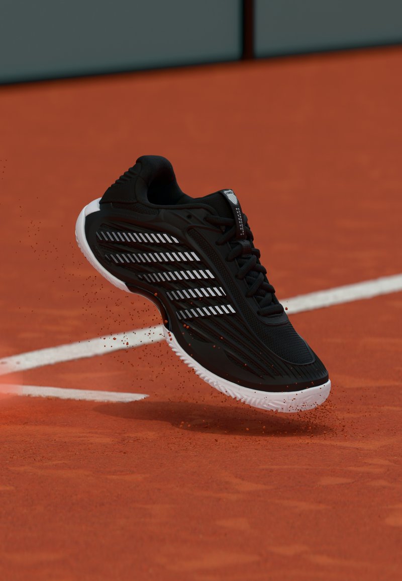 Μαύρο αθλητικό παπούτσι με λευκή σόλα και ριγωτό σχέδιο που σηκώνει κόκκινο πηλό σε γήπεδο τένις κοντά σε λευκές γραμμές.