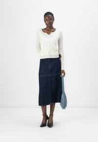Hvit ribbet cardigan, denim midi skjørt med sømdetaljer, blå håndveske og svarte høyhælte sko. Nøytral bakgrunn.