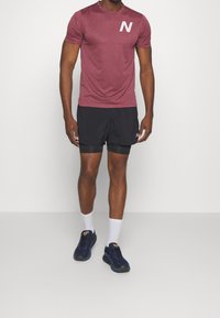 Maronenfarbenes T-Shirt mit kurzen Ärmeln für Sport, mit dem weißen "N"-Logo, schwarze Shorts mit integrierter Unterhose, weiße Socken und dunkelblaue Sneakers.
