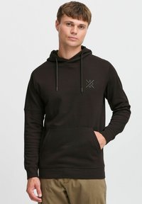 Schwarzer Kapuzenpullover aus einem weichen, strukturierten Stoff. Mit einer Fronttasche und einem subtilen Logo auf der Brust. Lange Ärmel mit gerippten Bündchen.