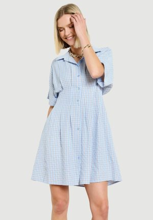 Frau lächelt, trägt ein locker sitzendes, kurzärmeliges, hellblau kariertes Kleid mit Kragen und Taschen, dazu goldene Halskette und Armband.