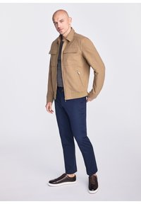 Jasny beżowy lekki kurtka z dwoma kieszeniami na klatce piersiowej, zamek błyskawiczny z przodu i ściągaczem na dolnym brzegu, zestawiona z granatowymi spodniami i ciemnymi trampkami. Minimalistyczny design.