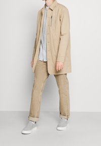 Beige košilové tričko s kapsou na zip, nošené přes světle šedé tričko, doplněné béžovými kalhotami a šedými slip-on botami.