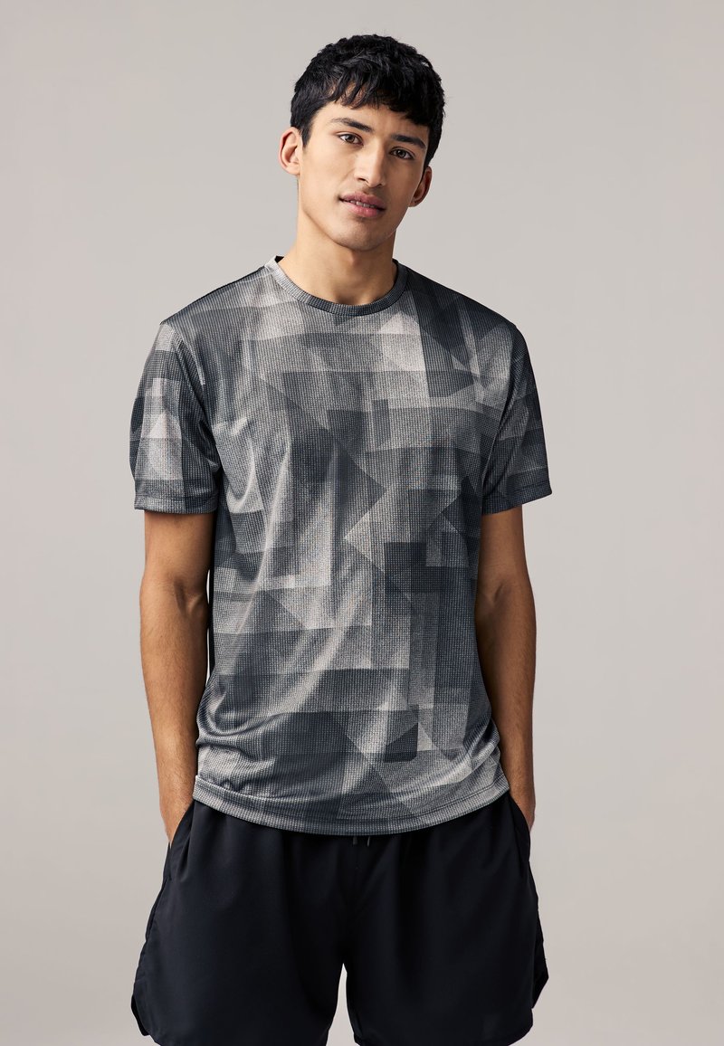 Graues Sport-T-Shirt mit geometrischem Muster, kurzen Ärmeln, rundem Ausschnitt und strukturiertem Finish. Kombiniert mit dunklen Shorts.