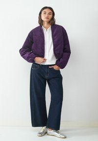 Lila Cordjacke im Bomberstil, weißes Hemd, dunkle weite Jeans und metallisch silberne Schuhe. Das Model steht vor einem weißen Hintergrund.