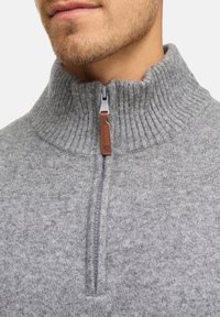 Grå stickad tröja med hög krage, med en halv dragkedja och en brun läderdragflik. Texturerad tyg med ribbade detaljer.