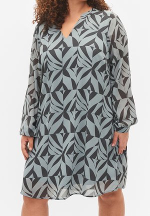 Kobieta ubrana w luźną sukienkę z długim rękawem w czarno-szary abstrakcyjny geometryczny wzór liści, z dekoltem w serek i przezroczystymi rękawami.