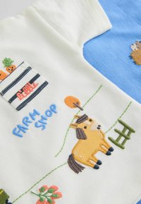 Morceaux de tissu blanc et bleu avec des éléments de ferme brodés, comprenant un cheval jaune, des carottes dans une caisse étiquetée "FARM SHOP" et des plantes.