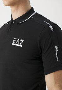 Homme portant un polo noir EA7 Emporio Armani avec un logo blanc sur la poitrine et un ruban de marque le long de l'épaule et de la manche.