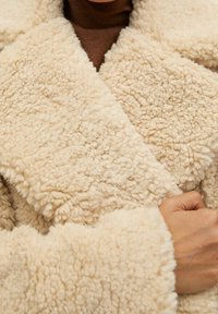 Kermanvärinen, pörröinen tekoturkkitakki, jossa on leveä kaulus ja päällekkäinen etuosa, sisältäen tekstuuria omaavaa kangasta ja löysät hihat.