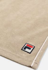 Coin d'une serviette en éponge beige avec un logo rectangulaire cousu en noir, rouge et blanc, et un bord ourlé rose clair.