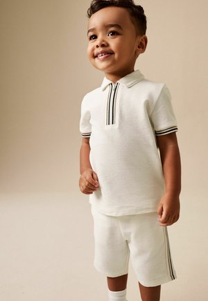 Väikelaps kannab valget lühikeste varrukatega polo särki ja sobivaid pükse mustade triipude detailidega, naeratades beeži tausta ees.