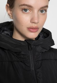 Doudoune noire avec un col montant, fermeture éclair sur le devant et tissu texturé. Le modèle porte un maquillage minimal et des boucles d'oreilles en strass.