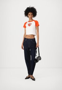 White crop top with orange raglan sleeves and heart graphic, paired with dark blue jeans. Black handbag and flats accent the outfit.