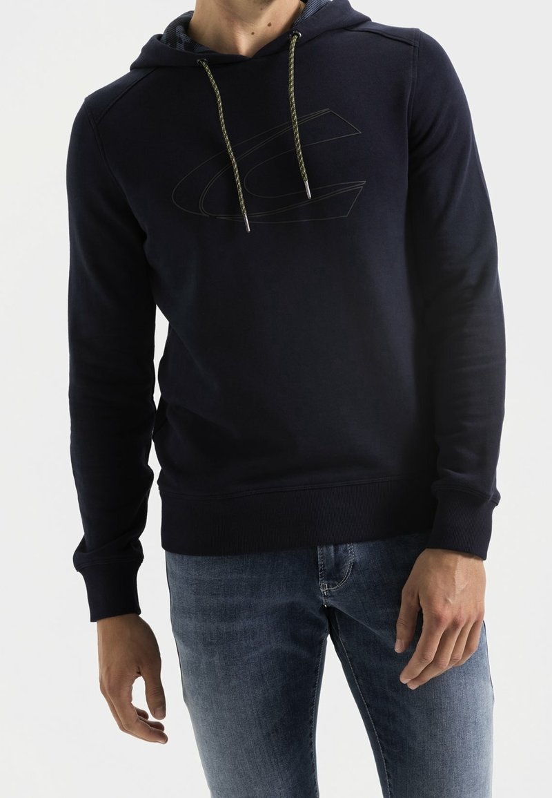 Felpa navy con cappuccio regolabile, caratterizzata da un grande logo in rilievo sul petto. Texture morbida, polsini e bordo a coste. Abbinata a jeans azzurri chiari.