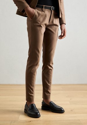 Person trägt braune, eng anliegende Hose, schwarzen Gürtel und schwarze Loafers, steht auf hellem Holzboden mit den Händen in der Nähe der Taschen.