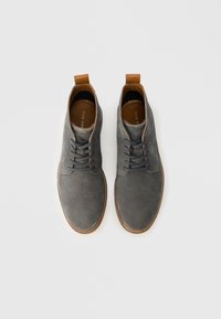 Szare zamszowe buty z brązowymi skórzanymi akcentami, okrągły czubek i sznurowany design. Posiadają kontrastującą podeszwę i przeszycia.