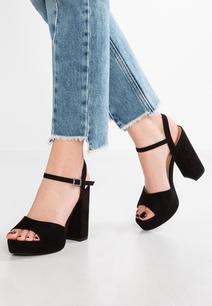 Benedenbenen met lichtblauwe spijkerbroeken met rafelige zoom en zwarte openhiel-sandalen met hoge hak en enkelbandjes, staand op een witte vloer.