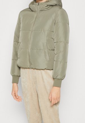 Veste matelassée vert olive clair avec capuche et fermeture éclair devant, portée avec un pantalon beige clair, montrant le haut du corps et les bras.
