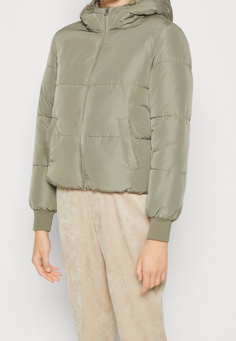 Veste matelassée vert olive clair avec capuche et fermeture éclair devant, portée avec un pantalon beige clair, montrant le haut du corps et les bras.