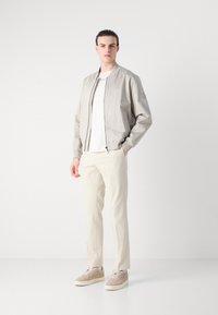 Lichtgrijze bomberjack met een ritssluiting, gecombineerd met een witte T-shirt en beige structuurbroek. Bruine suède sneakers maken de outfit compleet.