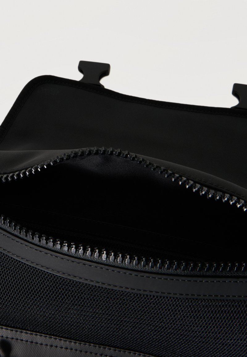 Poche d'un sac noir à fermeture éclair ouverte, montrant la doublure intérieure en filet et des attaches en plastique sur le bord supérieur, sur un fond uni.