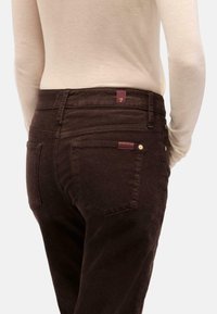 Pantaloni in corduroy marrone con vita alta, due tasche posteriori e un'etichetta color bordeaux. Texture morbida e motivo a righe verticali sono visibili.