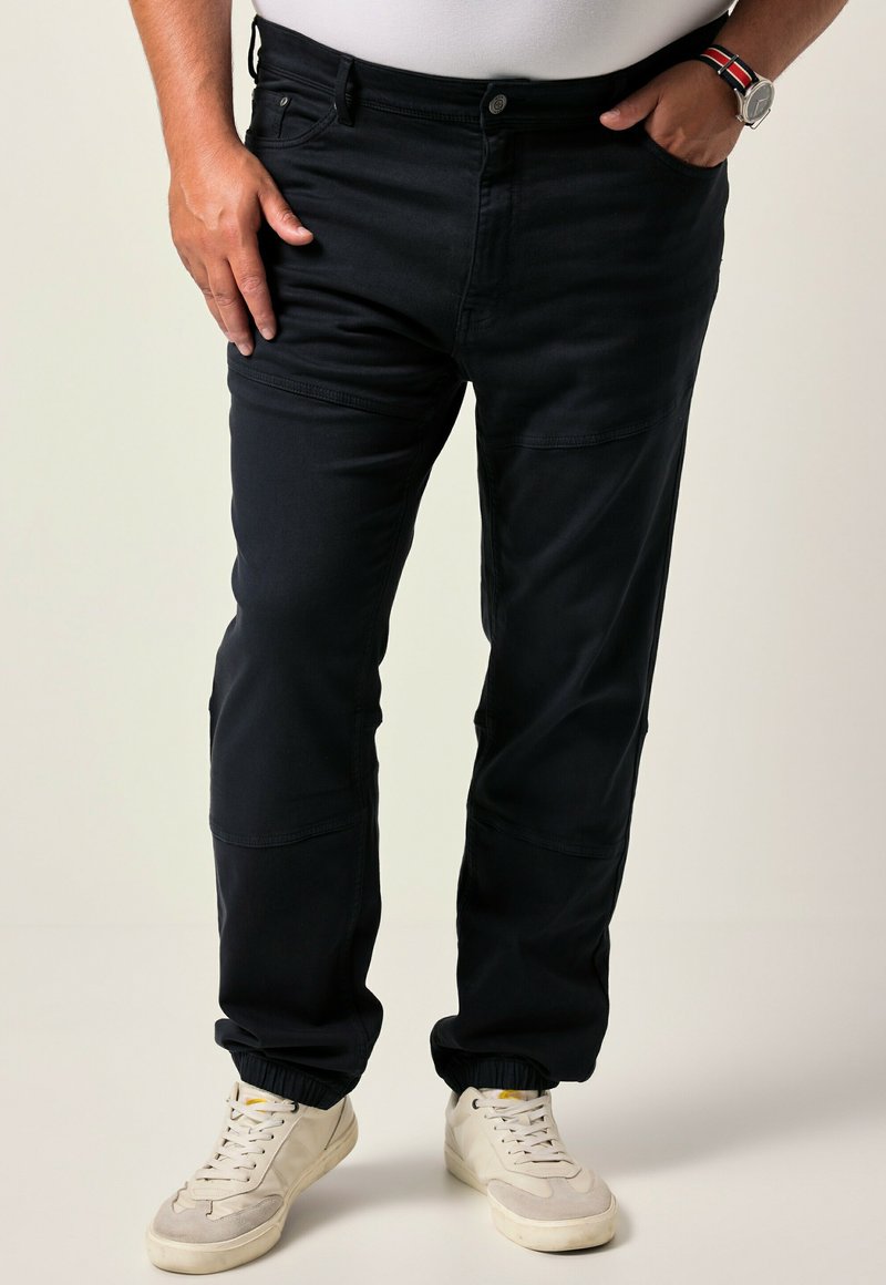Pantaloni blu scuro realizzati in un tessuto liscio, con un design affusolato e polsini elastici. Indossati con sneaker bianche e una camicia aderente sotto.