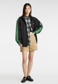 Chaqueta negra con mangas a cuadros verdes, camisa de cuadros debajo, pantalones cortos beige y zapatillas negras con tachuelas. Enfoque en la superposición casual y texturas.