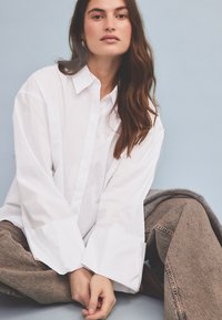Wite oversized button-up shirt met brede manchetten, gecombineerd met lichtbruine textuur pantalons. Model zit op de grond met een grijze trui in de buurt.