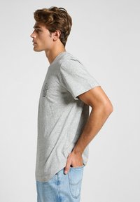 Camiseta de algodón gris con mangas cortas, cuello redondo y estampado gráfico negro en el pecho. Llevada con jeans de color azul claro.