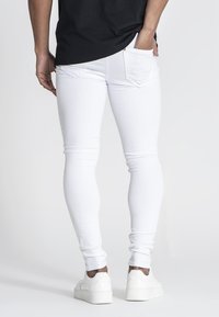 Jeans blancos ajustados con bolsillos traseros, combinados con una camiseta negra. La tela es suave, se ajusta al cuerpo, acompañados de zapatillas blancas.