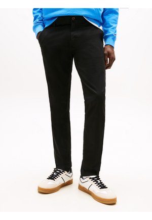 Persona vistiendo pantalones negros, zapatillas blancas con suelas de goma y una sudadera azul brillante con las manos en los bolsillos.