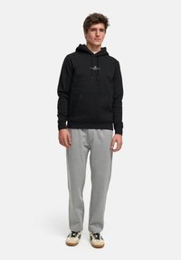 Svart hettegenser med lommepåse og hvit logo, kombinert med lysegrå joggebukser. Skoene har hvite og svarte farger.