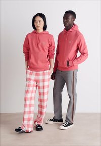 Δύο μοντέλα φοράνε ρούχα: μια ροζ φούτερ, καρό ροζ-άσπρο παντελόνι και γκρίζα τζιν με λωρίδα στο πλάι. Τα υποδήματα περιλαμβάνουν μαύρα και άσπρα αθλητικά παπούτσια.