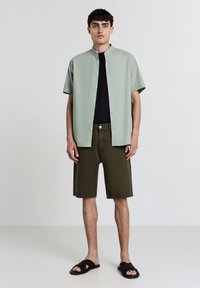 Kurzärmliges, hellgrünes Hemd mit Knöpfen und Stehkragen, kombiniert mit dunkelgrünen Shorts und schwarzen Sandalen. Glatter Stoff, lockere Passform.